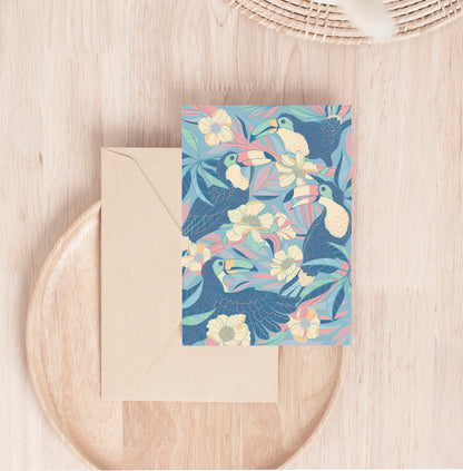 Colorful toucan and floral illustration on a card, set on a wooden surface with a beige envelope.