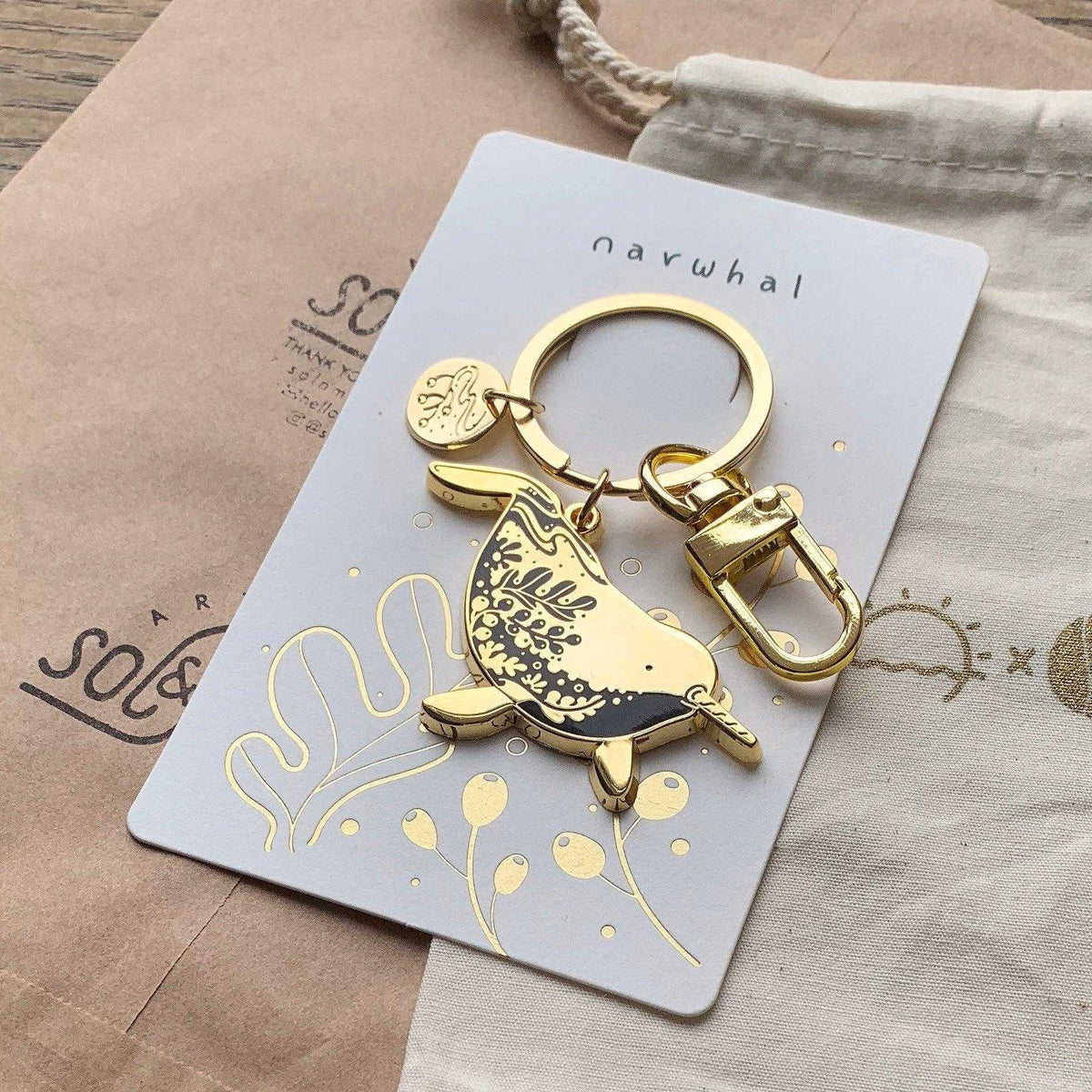 Golden narwhal enamel keychain on a decorative display card featuring line art and habitat illustration.