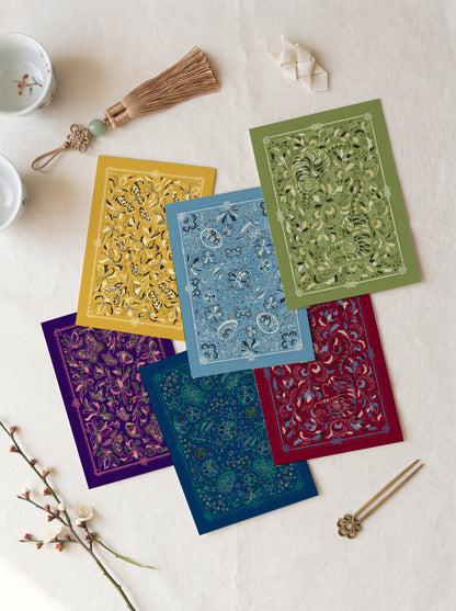 Six rectangular patterned cards or envelopes in yellow, green, blue, purple, teal, and red are arranged in an overlapping flat-lay on a light fabric surface, with small decorative props including a tassel, chopsticks, and tea cups nearby.
