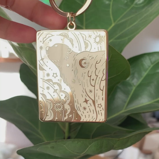 Gold-plated enamel keychain featuring a polar bear and northern sky design, attached to a green plant.