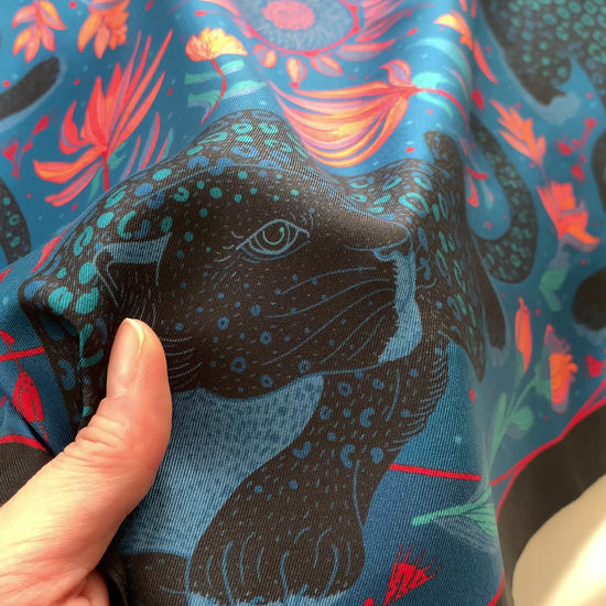 Hand holding a vibrant blue silk scarf featuring a leopard design with tropical flowers.