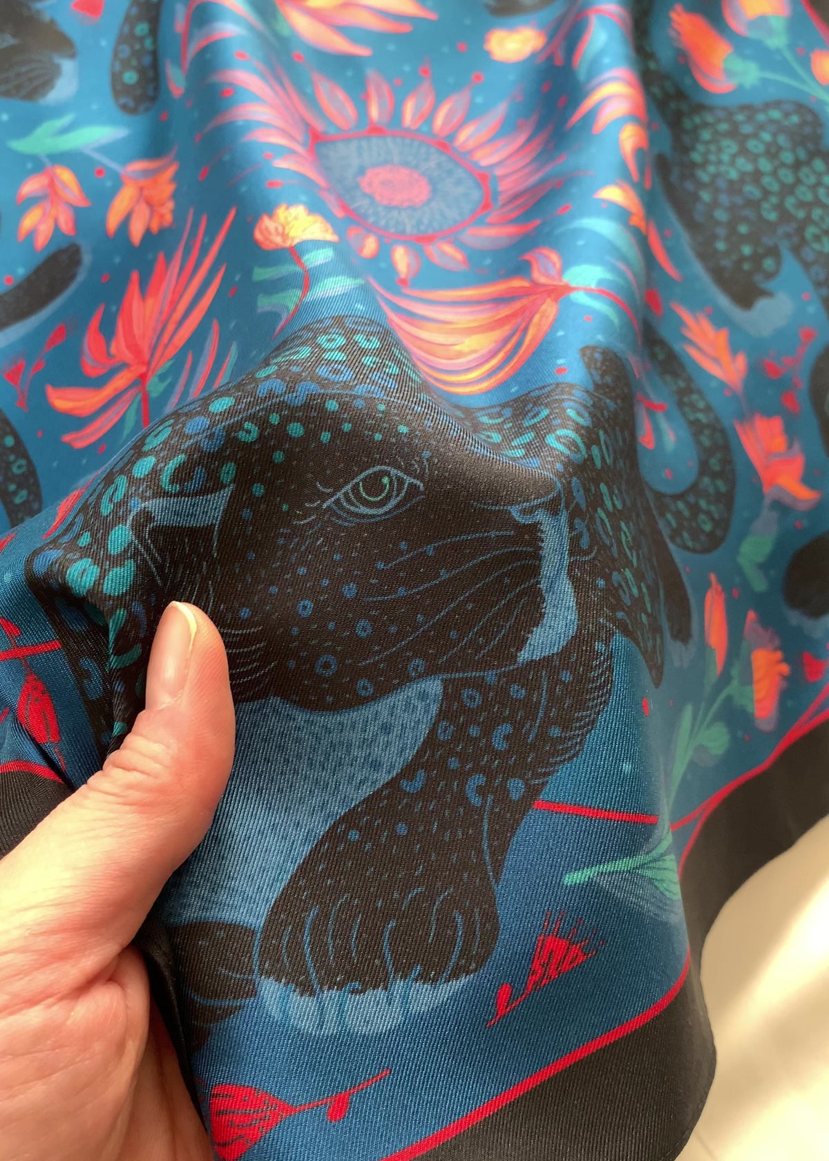 Hand holding a vibrant blue silk scarf featuring a leopard design with tropical flowers.