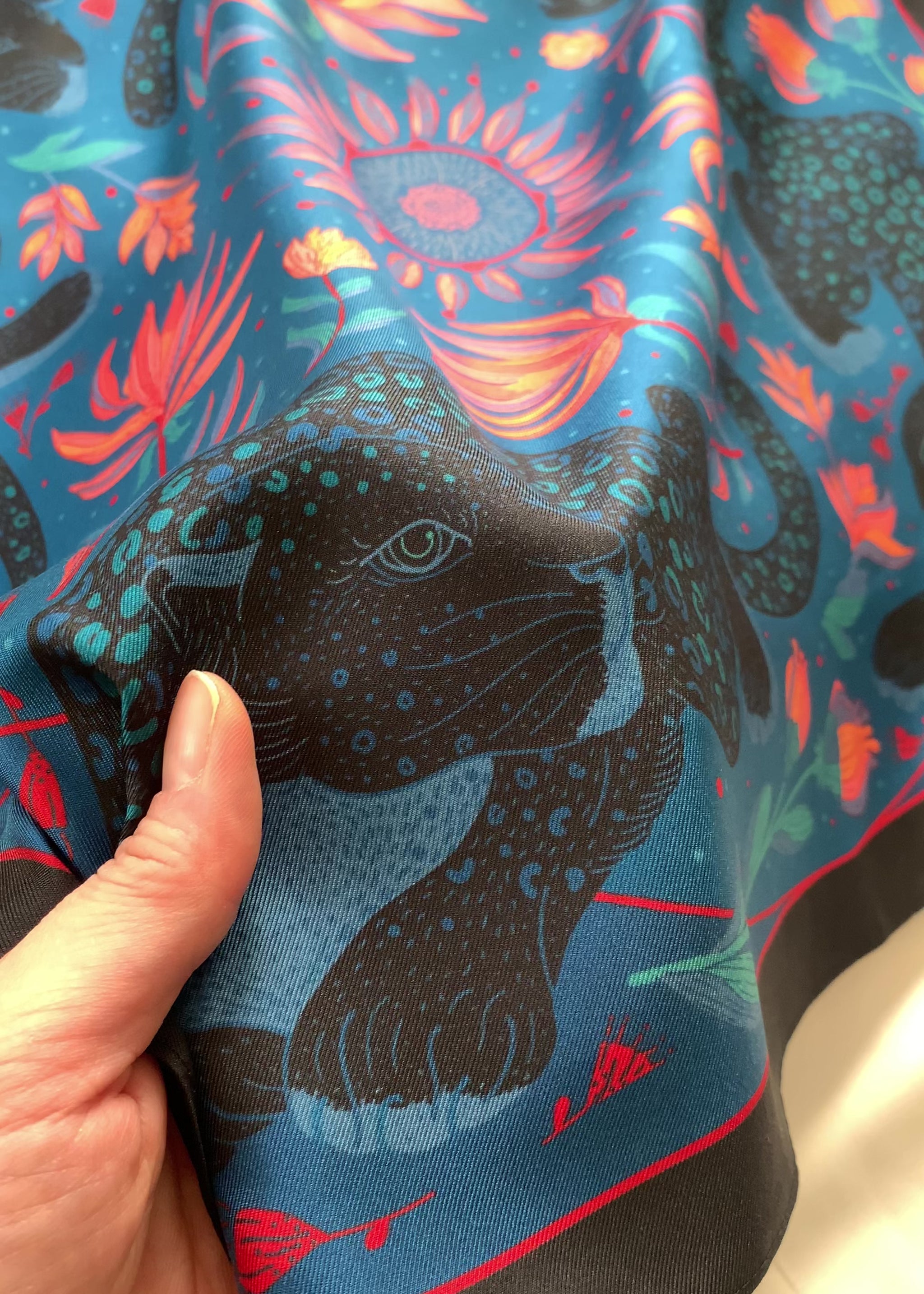 Hand holding a vibrant blue silk scarf featuring a leopard design with tropical flowers.