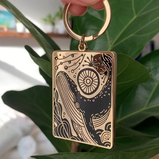Humpback whale and ocean enamel keychain hanging on a plant, featuring a gold-plated design.