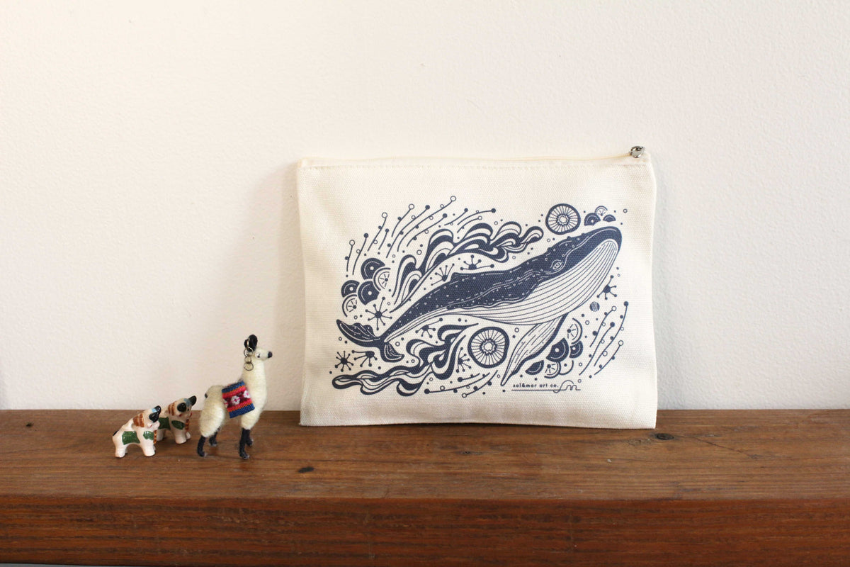 Canvas zipper pouch featuring whale art on a wooden surface with small figurines beside it.