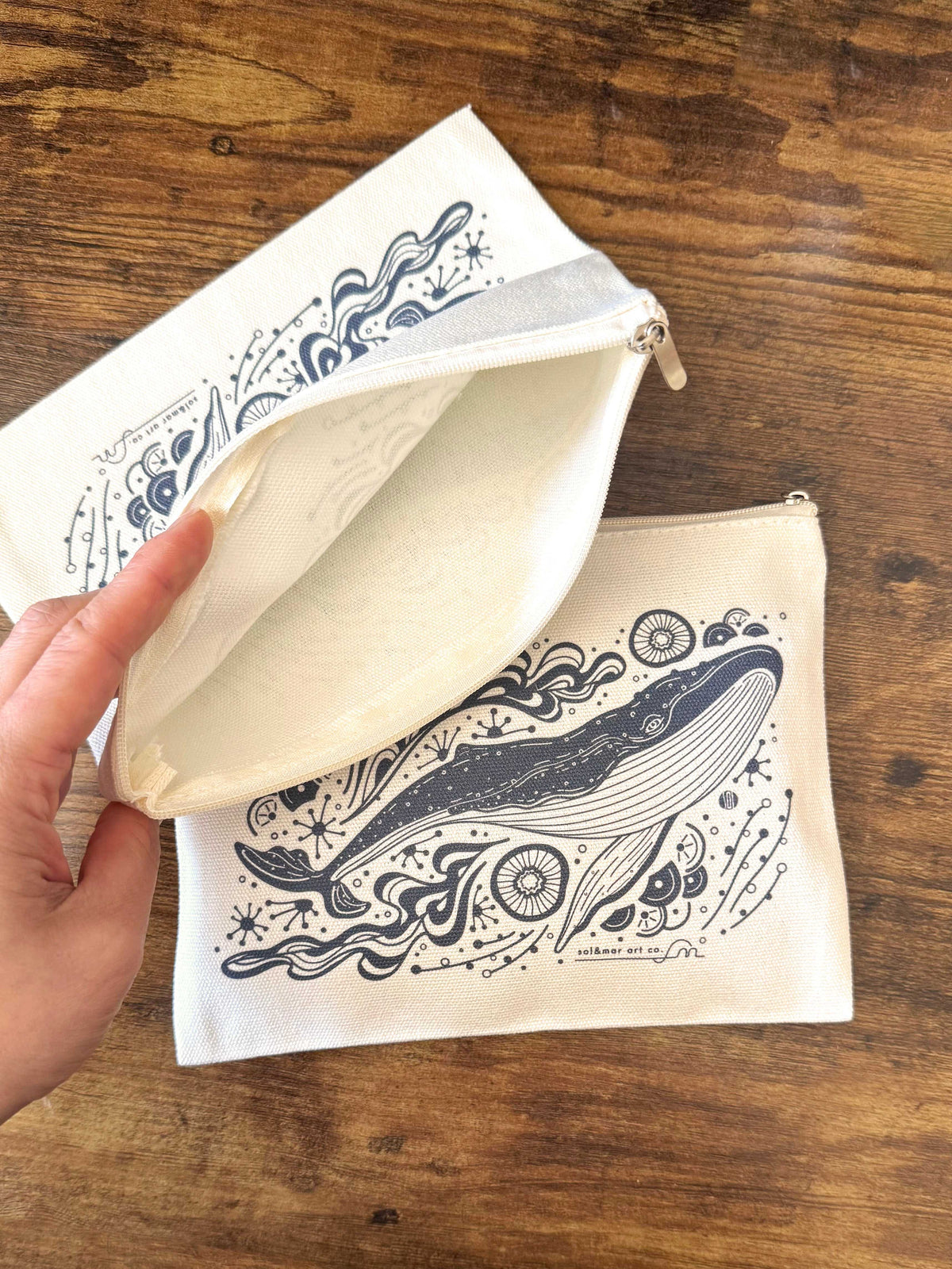 Canvas zipper pouch featuring whale illustration, perfect for makeup or storage, showing interior and design on wooden surface.