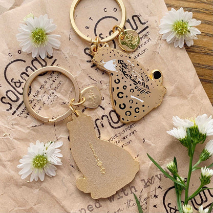 Amur Leopard enamel keychain with habitat illustration, adorned with delicate flowers and eco-friendly display card.