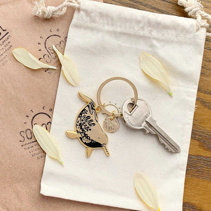 Narwhal enamel keychain in a drawstring bag, showcasing endangered species art and a circular charm, with a key.