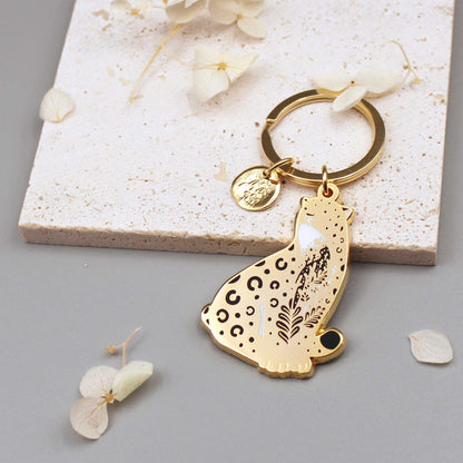 Amur Leopard enamel keychain with habitat design and gold charm on a stone background, promoting endangered animal awareness.
