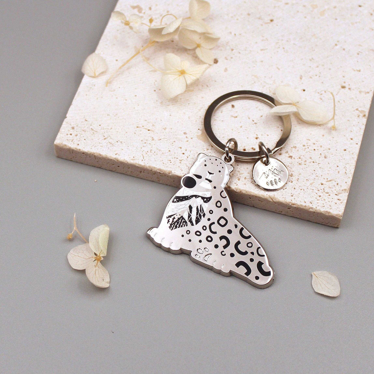 Snow Leopard enamel keychain featuring delicate art of its habitat on a round tag. Perfect for wildlife conservation advocates.
